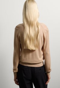 Jersey de punto beige de manga larga con puños y dobladillo acanalados, usado sobre pantalones negros. Textura suave, diseño minimalista y corte relajado.