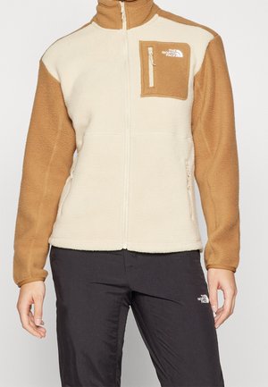 Person iført en beige og brun fleecejakke med lynlås og brystlomme samt sorte bukser, begge med The North Face-logo.