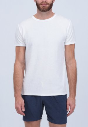 T-shirt blanc à col rond en coton, à manches courtes, texture lisse ; porté avec un short bleu foncé ; modèle debout sur un fond clair.