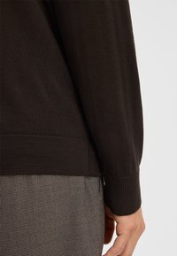 Brązowy sweter dzianinowy z prążkowanym dołem i mankietami. Tkanina ma gładką teksturę. Nosi się go na ubraniu w wzór houndstooth.
