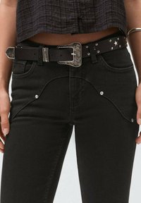 Vaqueros de mezclilla negra con una canesú de estilo western, detalles metálicos plateados y un cinturón negro con una hebilla decorativa plateada y tachuelas.