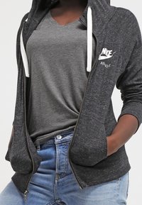Sudadera con cremallera de color gris oscuro con un acabado texturizado, cordones blancos y el logo de Nike. Lleva puesta sobre una camiseta gris de cuello en V, combinada con jeans claros.