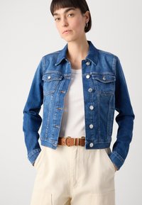 Giubbotto in denim blu con due tasche sul petto, bottoni argentati e cuciture a contrasto. Indossato sopra una camicia bianca e abbinato a pantaloni crema.