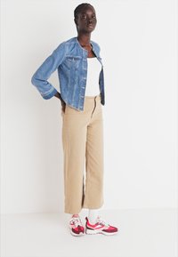 Jeans jakna, bela majica bez rukava, široke bež hlače i crvene patike. Manekenka pozira protiv jednostavne pozadine, prikazujući svakodnevnu odeću.