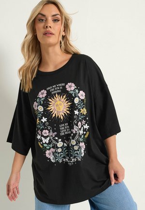 Blondynka nosi czarny oversize T-shirt z motywem słońca, księżyca, kwiatów, motyli i napisem: "Znajdź mnie tam, gdzie świeci słońce."