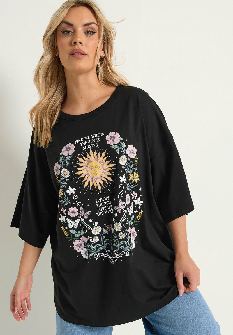 Femme blonde porte un T-shirt noir oversize avec soleil, lune, fleurs, papillons et texte : « Trouve-moi là où le soleil brille. »