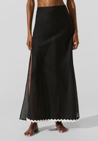 Kvinde iført en lang sort nederdel med et højt sideslid og hvid zigzagkant, stående barfodet mod en ensfarvet grå baggrund.