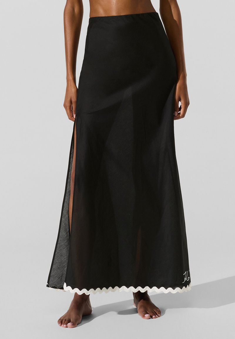 Kvinde iført en lang sort nederdel med et højt sideslid og hvid zigzagkant, stående barfodet mod en ensfarvet grå baggrund.