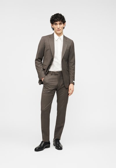 Καφέ κοστούμι με ραμμένη σχεδίαση, που περιλαμβάνει σακάκι με ένα κουμπί, πέτο με σχισμή και παντελόνι στενής γραμμής. Λευκό πουκάμισο από κάτω.