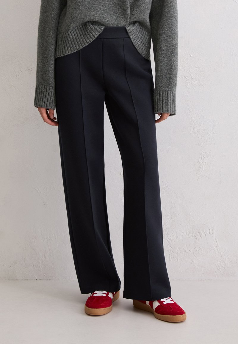 Granatowe spodnie o szerokich nogawkach z gładką teksturą i przednim szwem. Połączone z czerwonymi trampkami z białymi sznurówkami i beżową podeszwą.