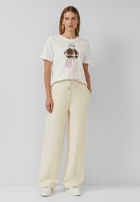 Frau in einem weißen T-Shirt mit einem grafischen Druck einer stilvollen Figur und cremefarbenen weiten Hosen, stehend vor einem schlichten grauen Hintergrund.