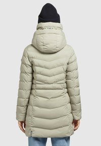 Lys beige pufferjakke med chevronmønster, der har en stor hætte og en ribbet sort strikhue. Glat tekstur med lange ærmer og lommer.