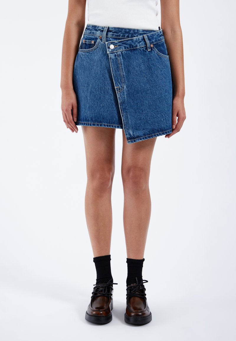 Jupe en jean mini avec un design légèrement asymétrique, couleur bleu clair, coutures visibles et poches avant ; assortie à des chaussures sombres et des chaussettes.