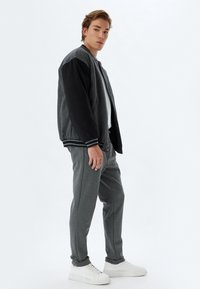 Giubbotto bomber nero e grigio con polsini a righe, indossato sopra una maglietta bianca, abbinato a pantaloni jogger grigi e sneakers bianche.
