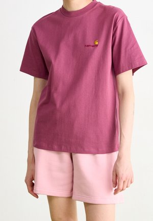 Persoon die een bordeauxrode Carhartt T-shirt met korte mouwen en lichtroze korte broek draagt, van nek tot halverwege de dijen, afgebeeld tegen een lichte achtergrond.