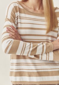 Bež ja valge triibuline sviiter, millel on lõdvestatud istuvus, lai kaelus ja soonikkoes manused. Pehme kudumine vahelduvate heleda ja tumeda triibuga.