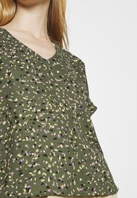 Blouse florale verte avec un col en V, ornée d'un imprimé éparpillé en violet, jaune et noir. Tissu léger et manches courtes.