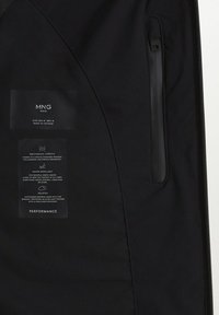 Veste imperméable noire avec stretch mécanique, dotée d'une poche zippée et d'étiquettes détaillant les matériaux et les instructions d'entretien.