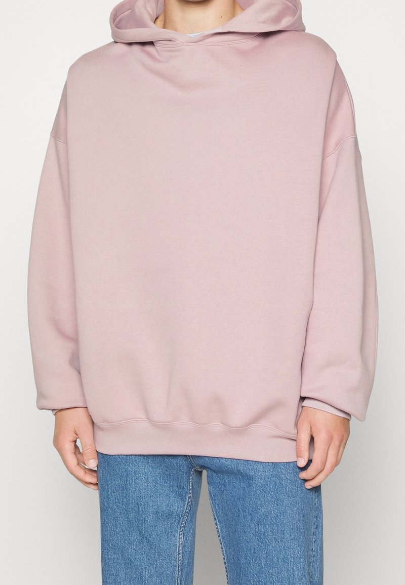 Personne portant un sweat à capuche rose clair oversize avec épaules tombantes et jean bleu, debout devant un fond uni.