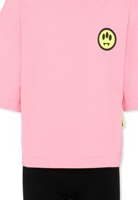 Camiseta rosa de manga corta con un pequeño logo de cara sonriente amarilla, que presenta ojos negros y colmillos, en la parte superior izquierda del pecho.