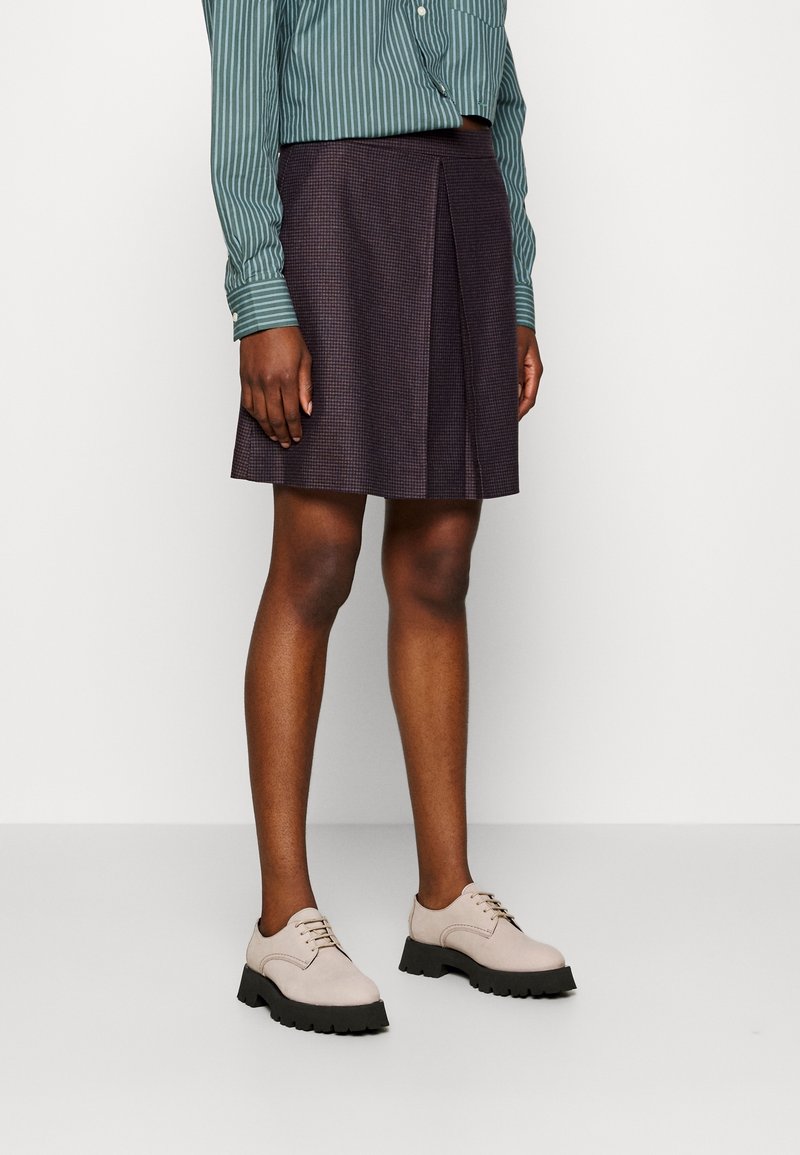 Gonna a pieghe a scacchi scuri abbinata a una camicia button-up a righe verde e scarpe lace-up rosa chiaro con suole nere.