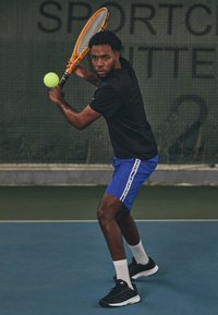 Τενίστας με μαύρο μπλουζάκι και μπλέ σορτς, κρατώντας μια πορτοκαλί ρακέτα με μια πράσινη μπάλα. Φορά μαύρα παπούτσια και λευκές κάλτσες.