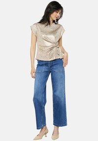 Top con stampa leopardata dorata con dettaglio annodato sul lato, abbinato a jeans larghi a vita alta blu e décolleté beige a punta.