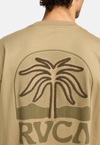 Beige katoenen t-shirt met een textuur van een palmboom in donkergroen, voorzien van een geperforeerde achtergrond, en de tekst "RVCA" onderaan.