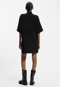 Zwarte oversized jurk met korte mouwen en een hoge hals, gemaakt van een zacht materiaal. Gestyled met hoge zwarte laarzen. Strak en minimalistisch design.