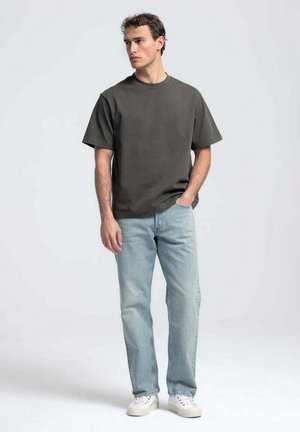 Man staat, draagt een donkergrijs oversized t-shirt, lichtblauwe jeans en witte sneakers, met hand in zak, kijkt naar de zijkant.