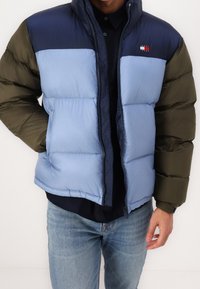 Prošívaná bunda s modrou a námořnicky modrou horní částí, zelenými rukávy, zipovým zapínáním a prošívaným vzorem. Nositelná přes tmavé tričko a džínové kalhoty.