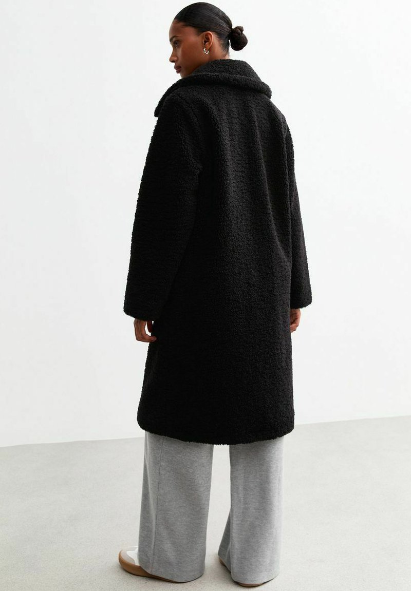 Czarny pluszowy płaszcz o fakturowanej powierzchni, z dużym kołnierzem i długimi rękawami, w zestawieniu z szarymi spodniami o szerokich nogawkach. Minimalistyczne tło.
