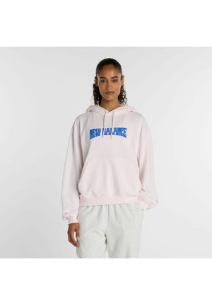 Frau steht und trägt einen hellrosa New Balance Hoodie und hellgraue Jogginghosen vor einem schlichten weißen Hintergrund.