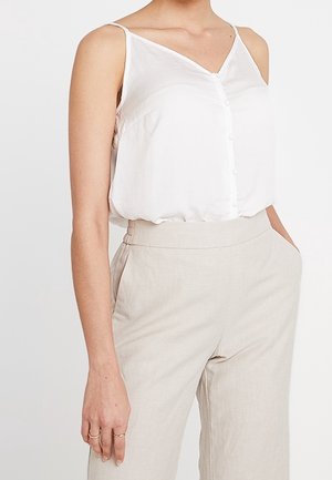 Frau trägt ein ärmelloses weißes, knopfbewehrtes Oberteil und hoch taillierte beige Hosen, steht mit einer Hand in der Tasche vor einem schlichten Hintergrund.