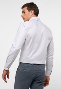 Hellgraues Langarmhemd mit einem gepflegten Kragen und knöpfbaren Manschetten, das eine subtile Textur aufweist. Getragen mit dunkelgrauen Hosen.