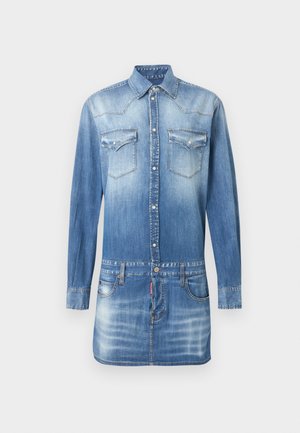 Γυναίκα με ξεθωριασμένο μπλε τζιν πουκαμισοφόρεμα με τσέπες στο στήθος και λεπτομέρειες με κουμπιά, φοράει μαύρες ανοιχτές ψηλοτάκουνες γόβες, στεκόμενη μπροστά σε λευκό φόντο.