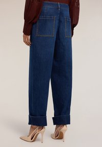 Pantalones de mezclilla anchos de color azul oscuro con costuras laterales visibles, puños enrollados y dos bolsillos traseros con detalles de remaches.