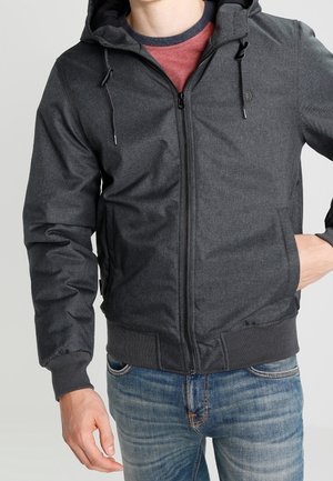 Sudadera gris con cremallera y capucha, con una textura suave, puños elásticos y una cintura ajustable. Se lleva encima de una camiseta roja y jeans azules.