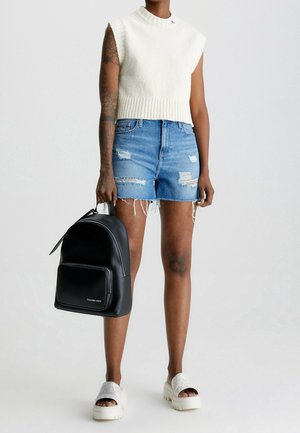 Person iført hvid ærmeløs striktop, slidte blå denimshorts, hvide platåsandaler og holder en sort rygsæk i venstre hånd.