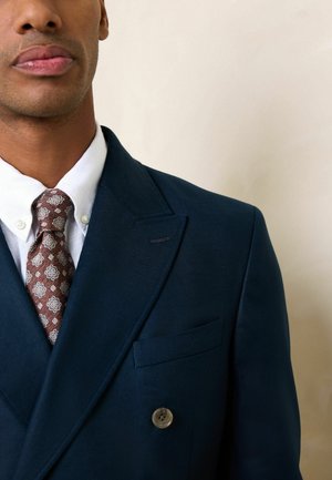 Mann trägt einen marineblauen Doppelreiher-Anzug, weißes Hemd mit Knöpfen und gemusterte braune Krawatte vor neutralem Hintergrund.