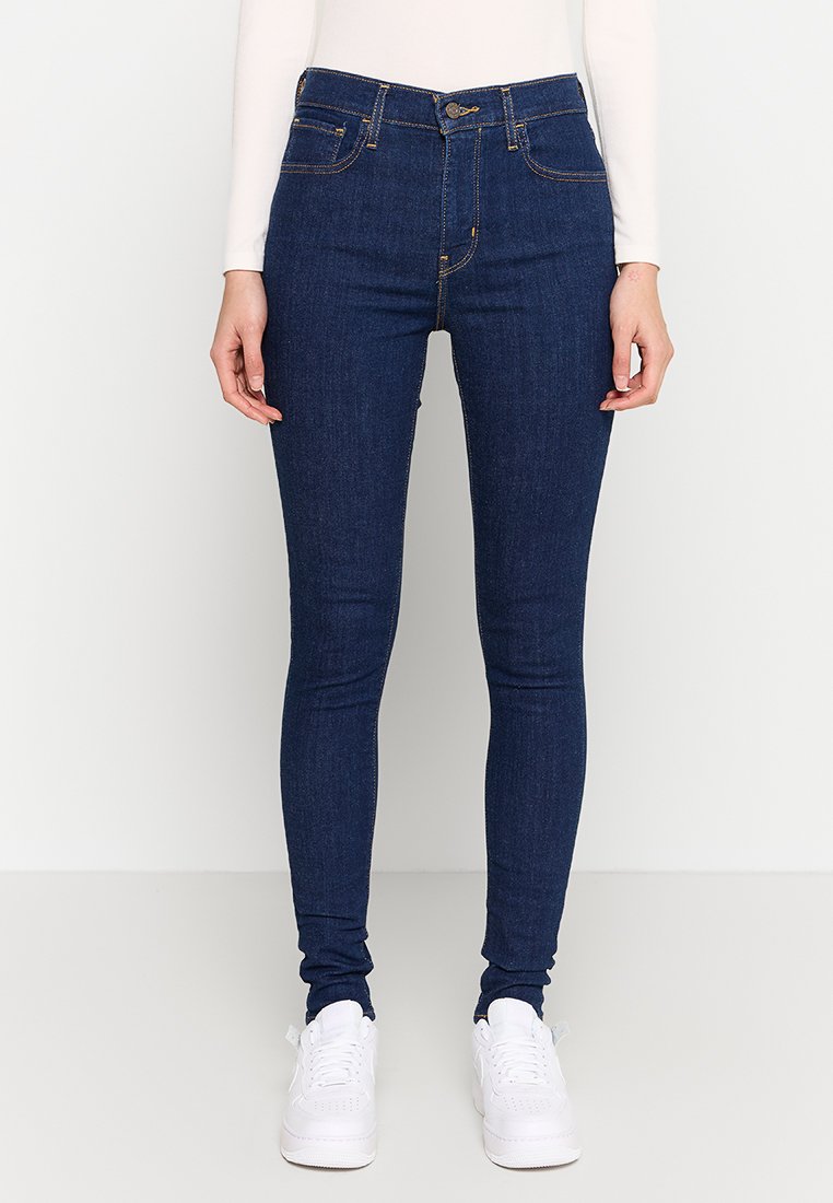 Marineblaue Slim-Fit-Jeans aus Denim-Stoff mit Fünf-Taschen-Design, Knopfverschluss und Kontrastnähten; kombiniert mit weißen Sneakers.