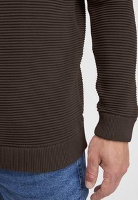 Primo piano del braccio e della mano di una persona che indossa un maglione a coste scuro a maniche lunghe e jeans blu su uno sfondo semplice.