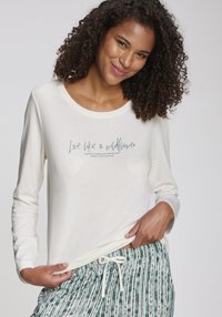 Chemise beige à manches longues avec encolure arrondie, portant le texte bleu "Vis comme une fleur sauvage." Associée avec un pantalon vert à cordon de serrage à motifs.