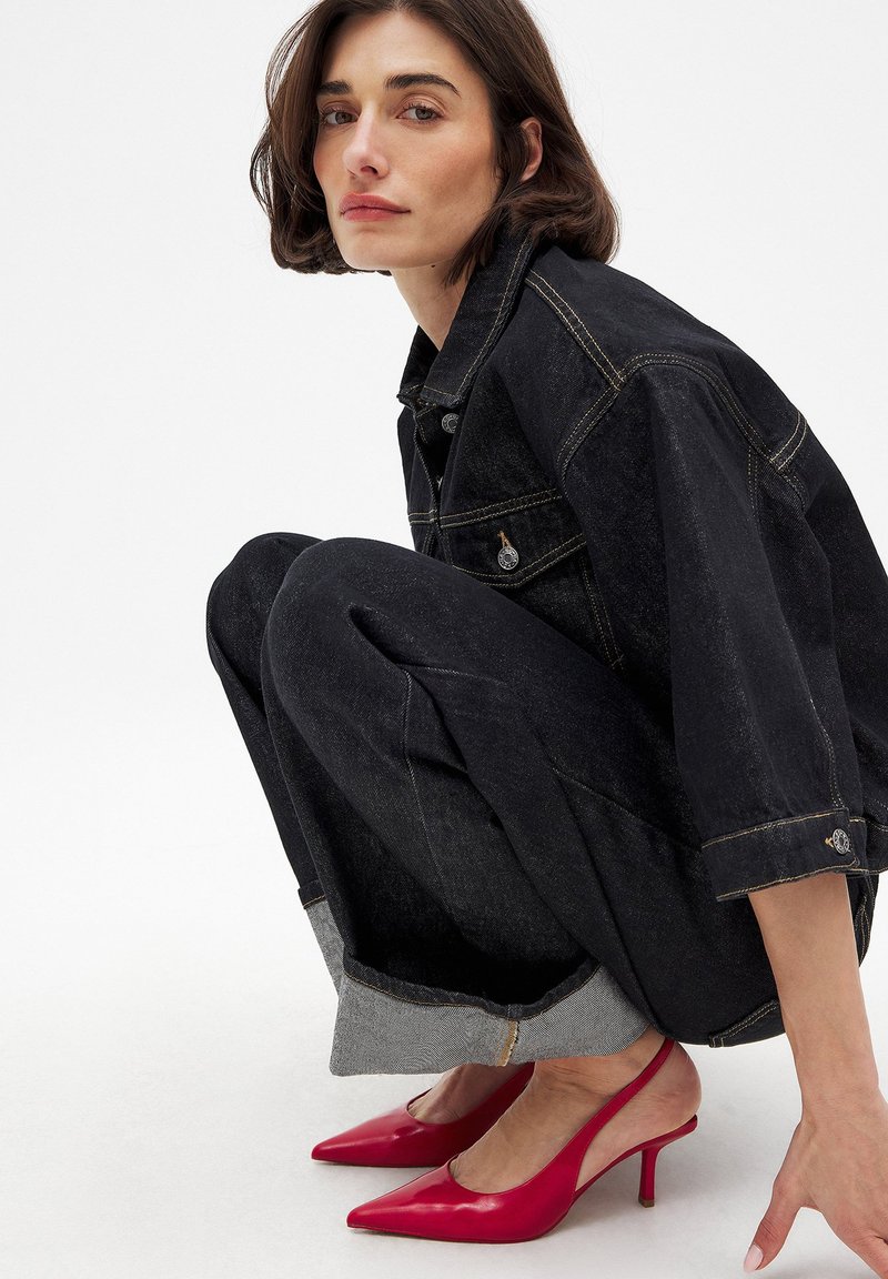 Veste en denim noir surdimensionnée et pantalon avec revers ; escarpins rouges à bout pointu. Texture lisse, détails de couture contrastants.