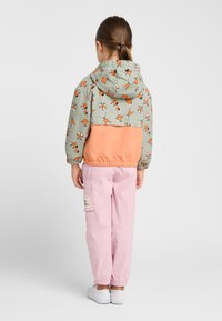 Veste coupe-vent colorée avec un haut à motifs verts comportant des oiseaux oranges, et une partie inférieure corail. Associée à un pantalon cargo rose clair.