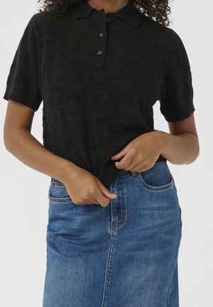 Donna che indossa una polo nera a maniche corte con texture e una gonna di jeans blu, che sistema l'orlo della maglietta con le mani.