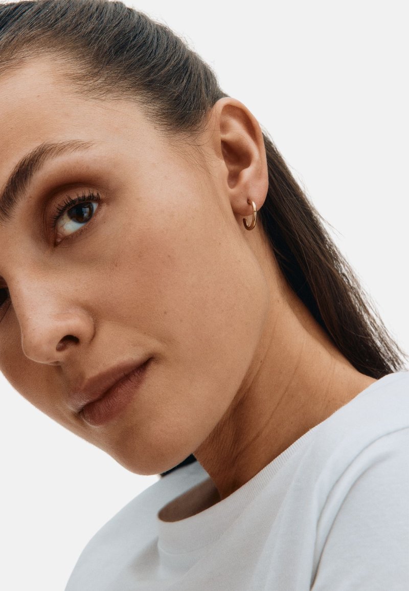 Kultainen rengaskorvakoru mallin korvassa, jossa on sileä, kiillotettu pinta ja pyöreä muoto neutraalia taustaa vasten.