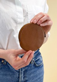 Apaļa brūna ādas maisiņš ar teksturētu virsmu un rāvējslēdzēju, turēts starp pirkstiem pret gaismas fonu un džinsiem.