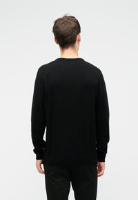 Jeune homme debout de dos, portant un pull noir à manches longues et un pantalon noir, sur un fond clair uni.