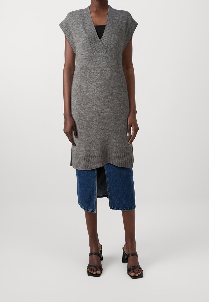 Femme portant une tunique en maille grise sans manches sur une jupe midi en denim bleu avec une fente à l'avant et des sandales noires à talons.